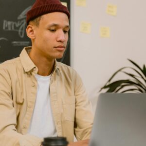 Young adult working on a laptop wearing a beanie in an office setting with notes on a chalkboard.
