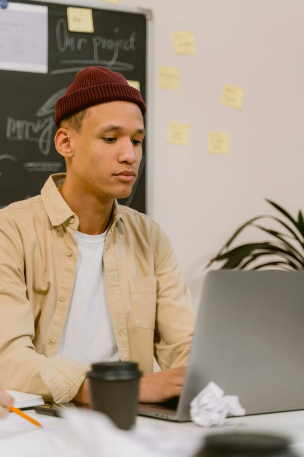 Young adult working on a laptop wearing a beanie in an office setting with notes on a chalkboard.